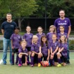 Junior Chefs A youth soccer team in purple jerseys poses with their coaches on a turf field, with one player kneeling in front of the ball.