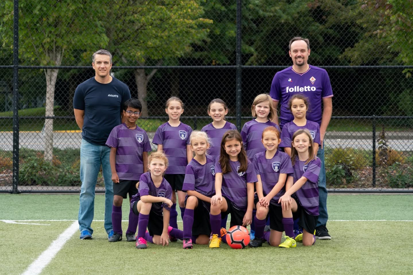 Alicorn Team Pictures A youth soccer team in purple jerseys poses with their coaches on a turf field, with one player kneeling in front of the ball.