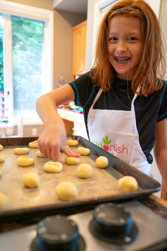 A young person in a black T-shirt and apron is placing small balls of dough onto a baking sheet.