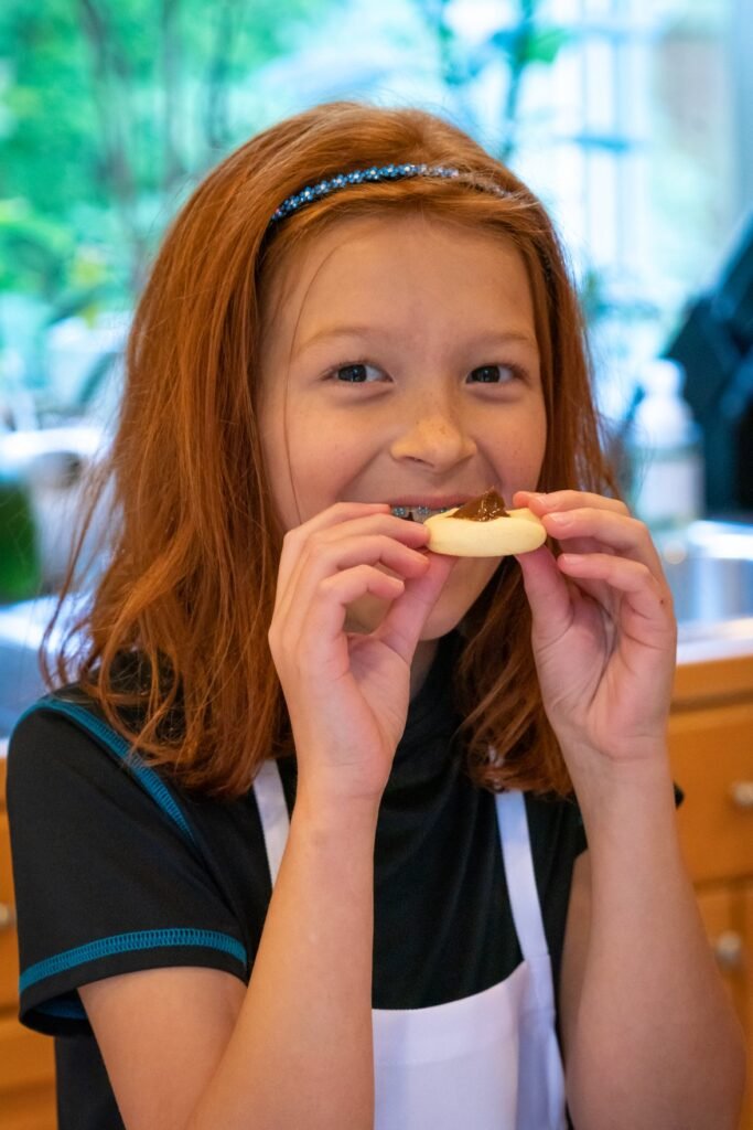 A young child wearing a headband and apron smiles joyfully while tasting a homemade pastry.