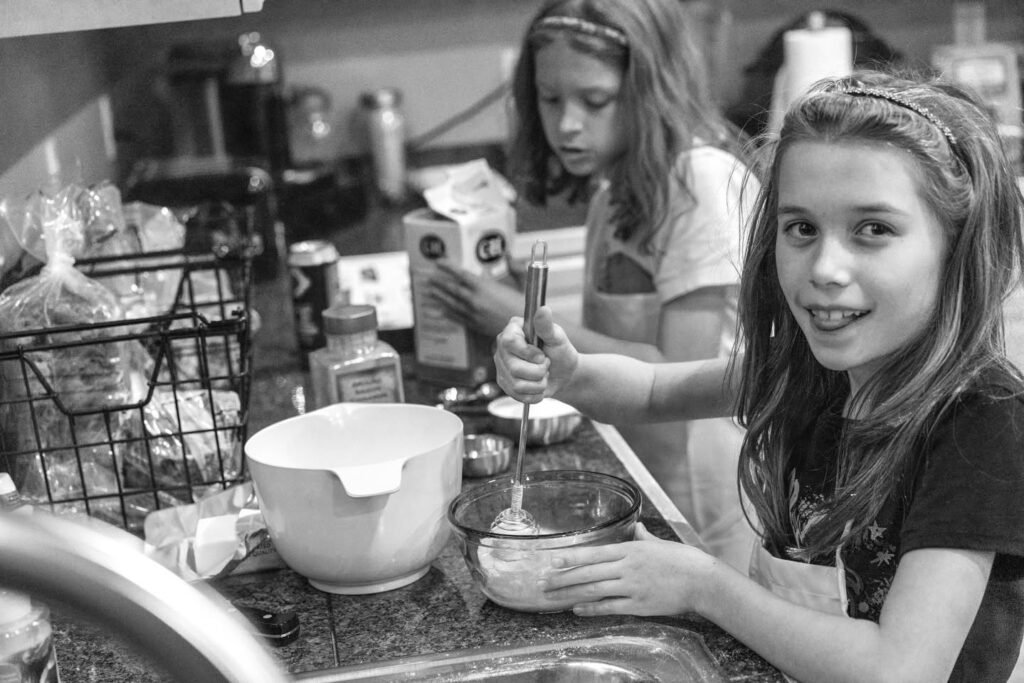 Two children standing together at the kitchen counter while one whisks. They are surrounded by supplies including a mixing bowl and utensils.