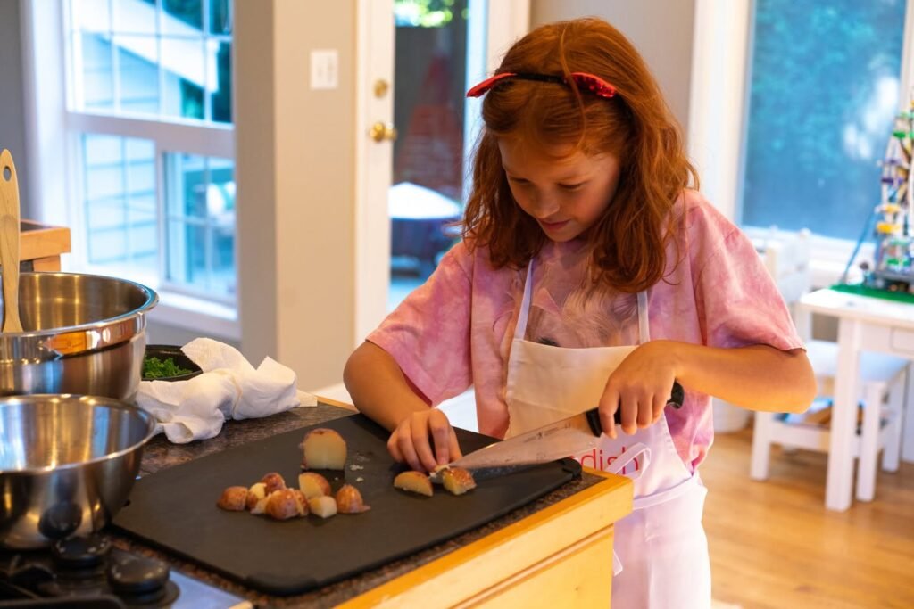 A child in an apron carefully cuts vegetables on a cutting board in a bright, sun-filled kitchen.