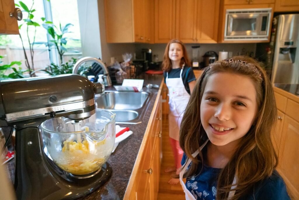 Two children smile happily while using a stand mixer to bake in a warm, wooden kitchen setting.