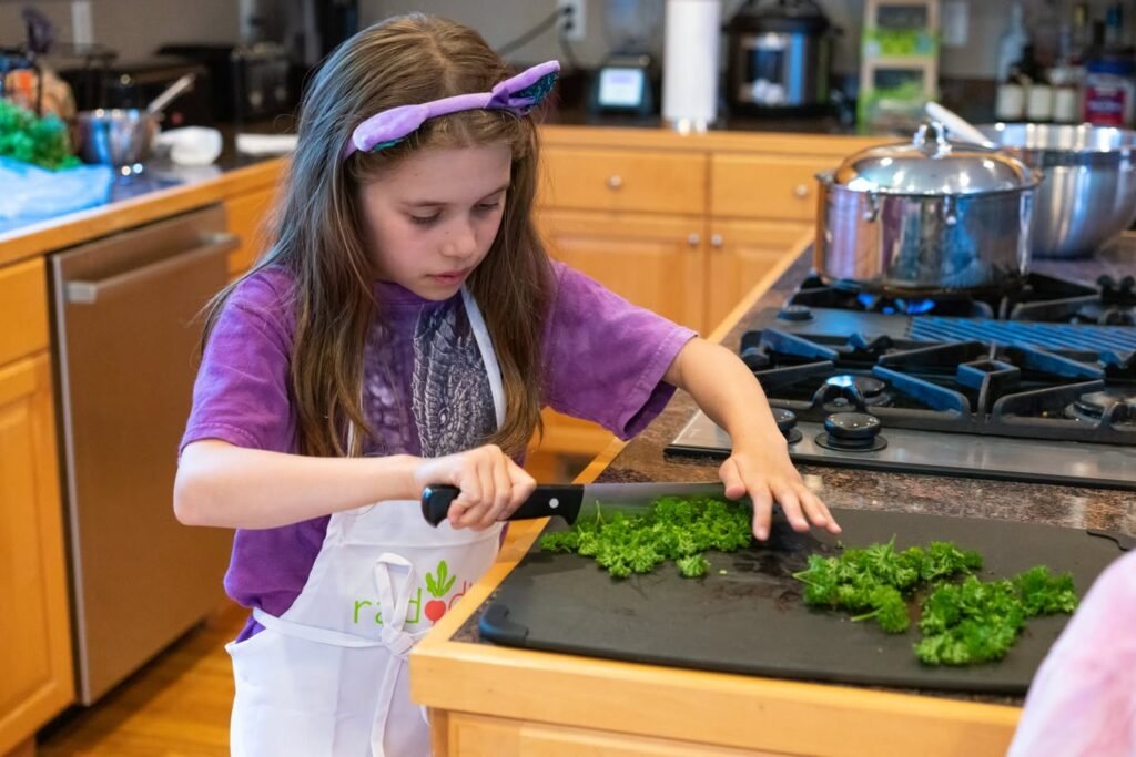 A child in a purple shirt and apron is chopping green herbs on a black cutting board in a bright kitchen with wooden cabinets and stainless steel appliances.