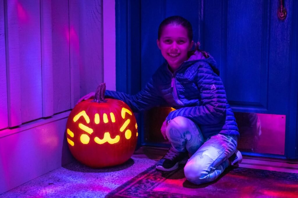 A child crouching beside a glowing jack-o'-lantern on a front porch under vibrant purple and pink lighting.