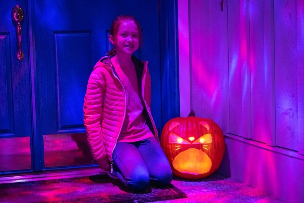 A person crouches beside a glowing jack-o'-lantern in a portrait orientation, illuminated by purple and blue lights on a front porch. An extreme close-up reveals the vertical texture of the door in bright neon hues.