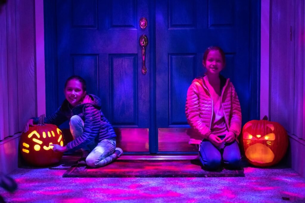 Two children pose in front of a dark front door lit by a carved pumpkin and purple mood lighting.