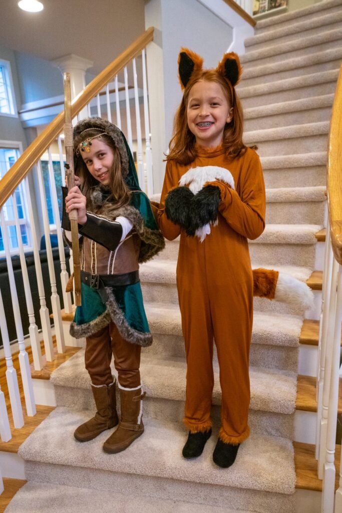 Two children dressed in playful Halloween costumes stand on a carpeted staircase, one in an animal onesie and the other in a fantasy outfit.