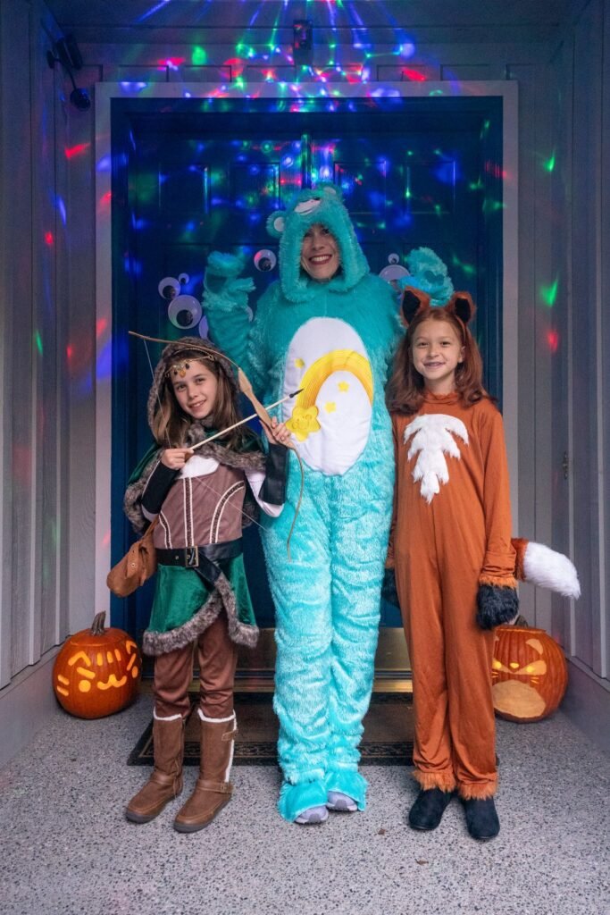 Three children in Halloween costumes, including a dragon and a fox, stand on a porch with festive lights and a carved pumpkin.