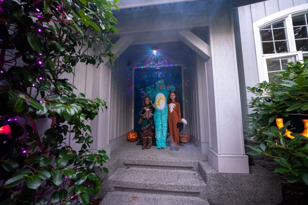 A festive Halloween display on a home porch with costumed figures, pumpkins, and glowing lights surrounded by lush greenery.