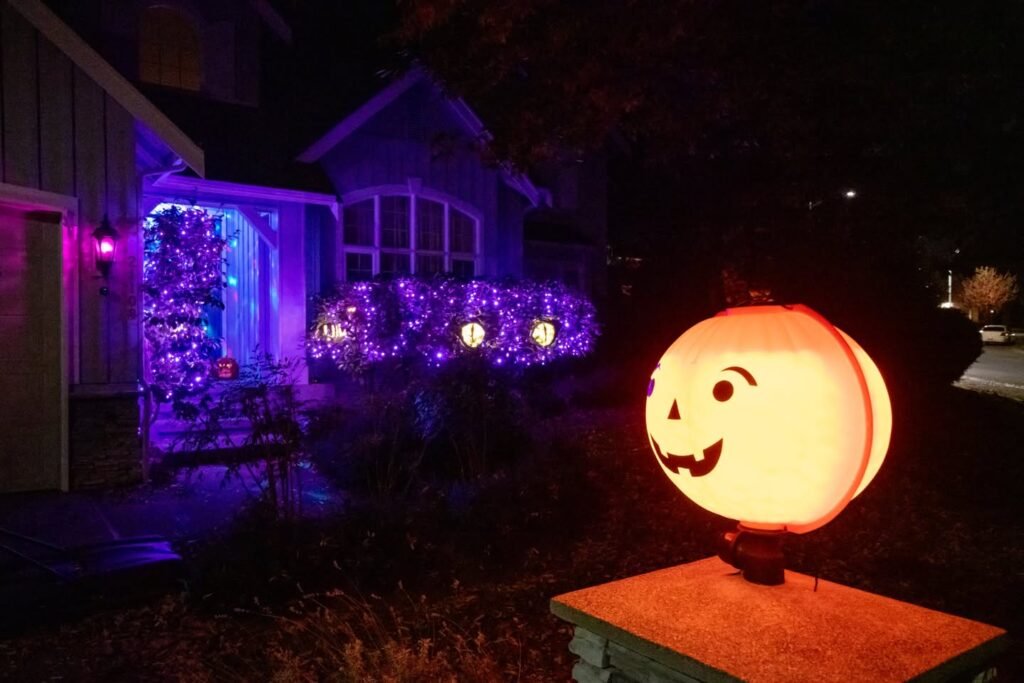 A large illuminated jack-o-lantern sits on a sidewalk in front of a house decorated with purple holiday lights.