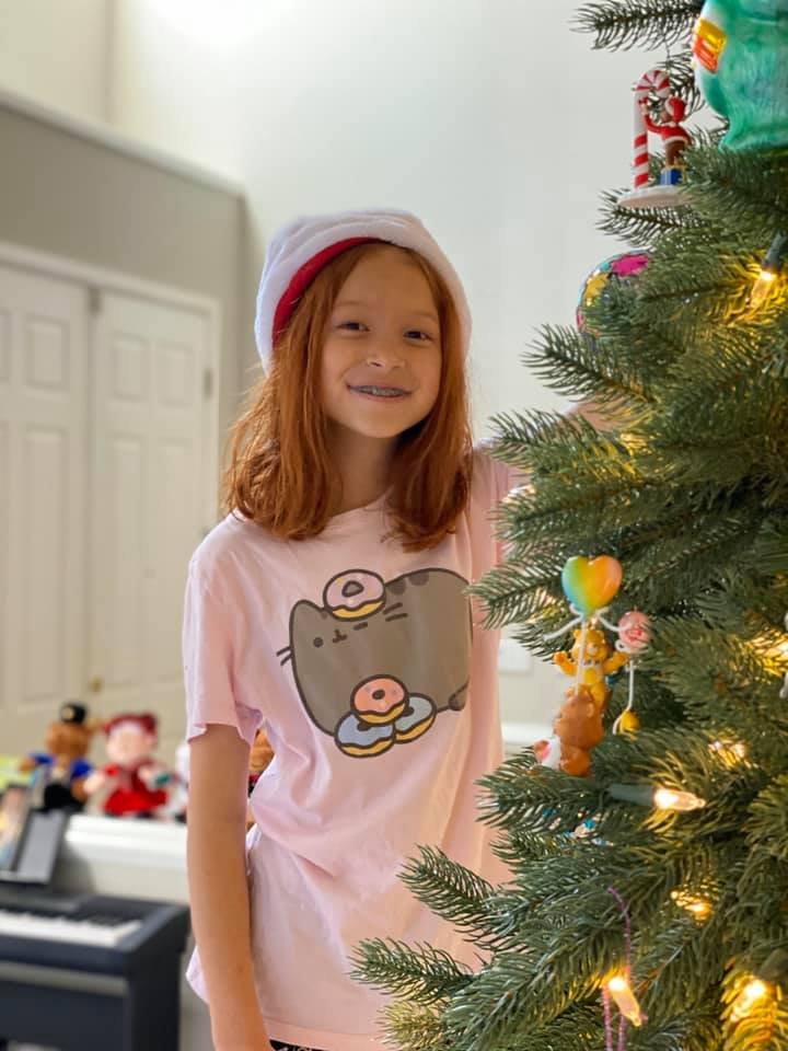 A young girl wearing a Santa hat and a pink cat-themed shirt standing next to a decorated Christmas tree with lights.
