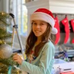 A girl wearing a red Christmas hat smiles while decorating the tree. Red stockings hang from the fireplace mantel in the background.