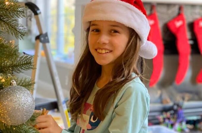 A girl wearing a red Christmas hat smiles while decorating the tree. Red stockings hang from the fireplace mantel in the background.