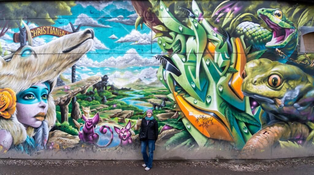 A person stands in front of a colorful mural in Portland, featuring fantasy characters like frogs and birds.