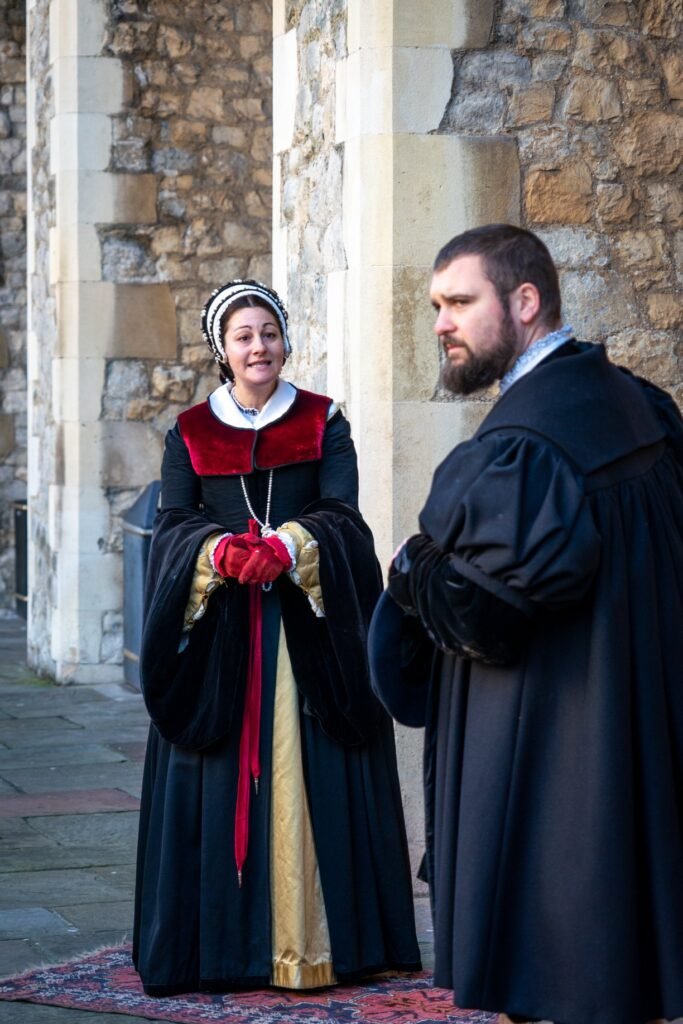 Two people in period Tudor-style Renaissance clothing standing on a stone patio. Detailed view of a black gown with a red velvet collar, gold trim, and red gloves.
