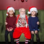 Copenhagen Santa Claus poses on a green couch with two girls wearing red Christmas hats. A festive holiday wreath hangs on the red wall behind them.