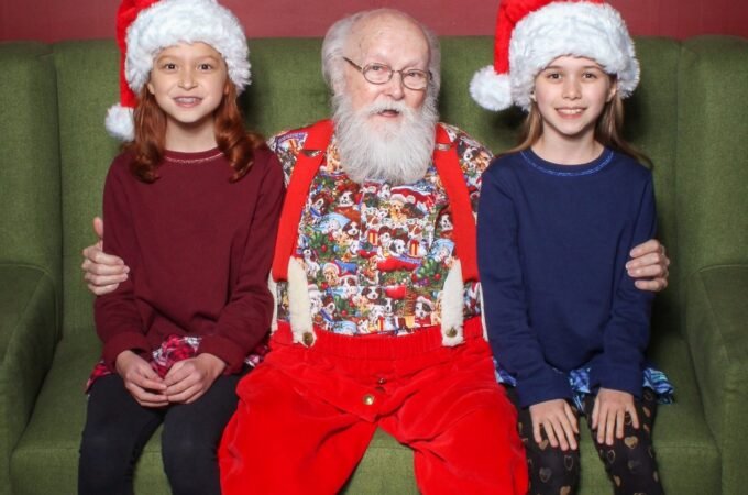 Santa Claus poses on a green couch with two girls wearing red Christmas hats. A festive holiday wreath hangs on the red wall behind them.