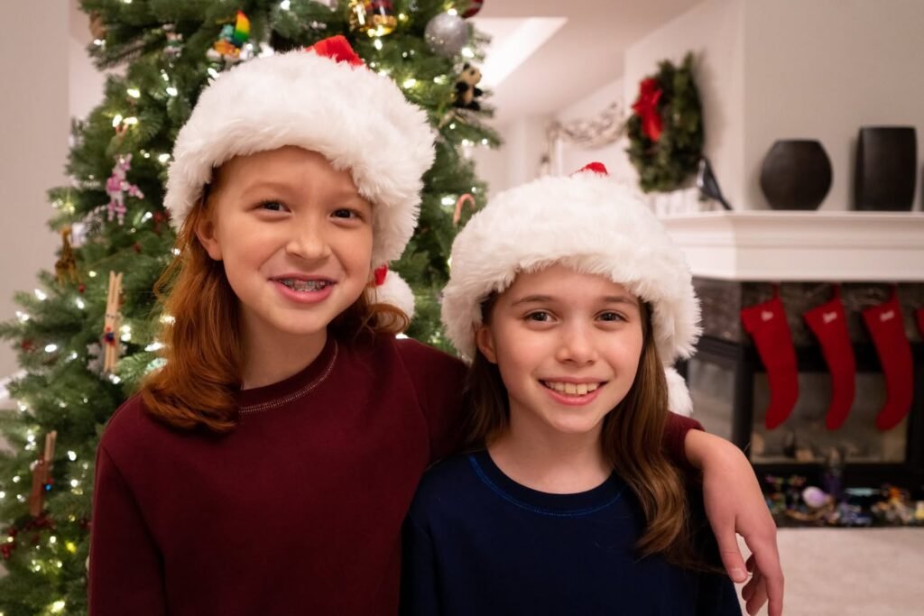 Two children wearing Santa hats smile in front of a Christmas tree. A decorated mantel with red stockings is visible in the background.