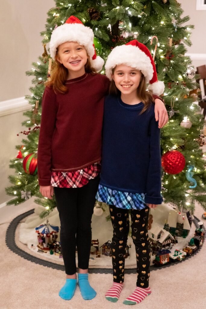 Two girls wearing Santa hats posing together in front of a decorated Christmas tree with festive lights and ornaments.