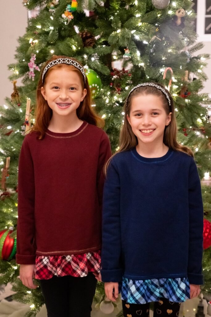 Two children in festive sweaters smiling in front of a decorated Christmas tree with colorful lights and ornaments.