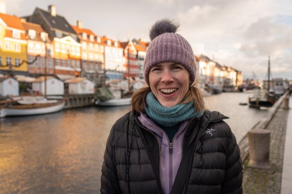 A person wearing a winter beanie smiles in front of the colorful Nyhavn waterfront in Copenhagen, Denmark.