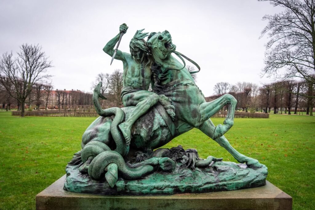 A verdigris bronze statue depicting a knight battling a dragon stands on a green grassy field in a park.