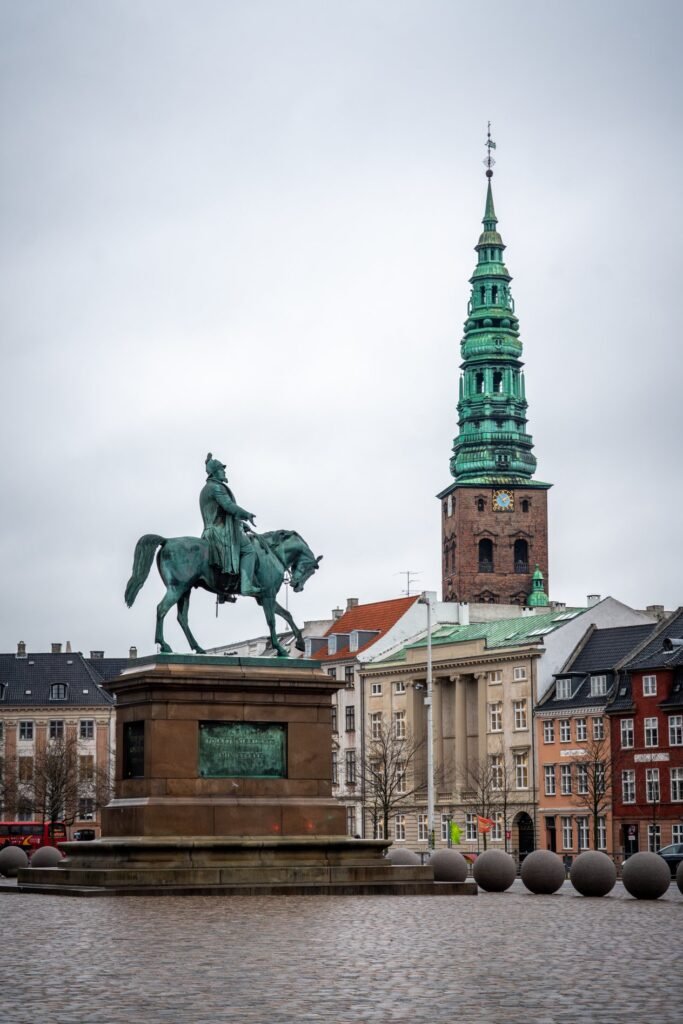 A bronze equestrian statue stands prominently on a stone pedestal in a European town square, with an ancient church spire and colorful historic buildings in the background.