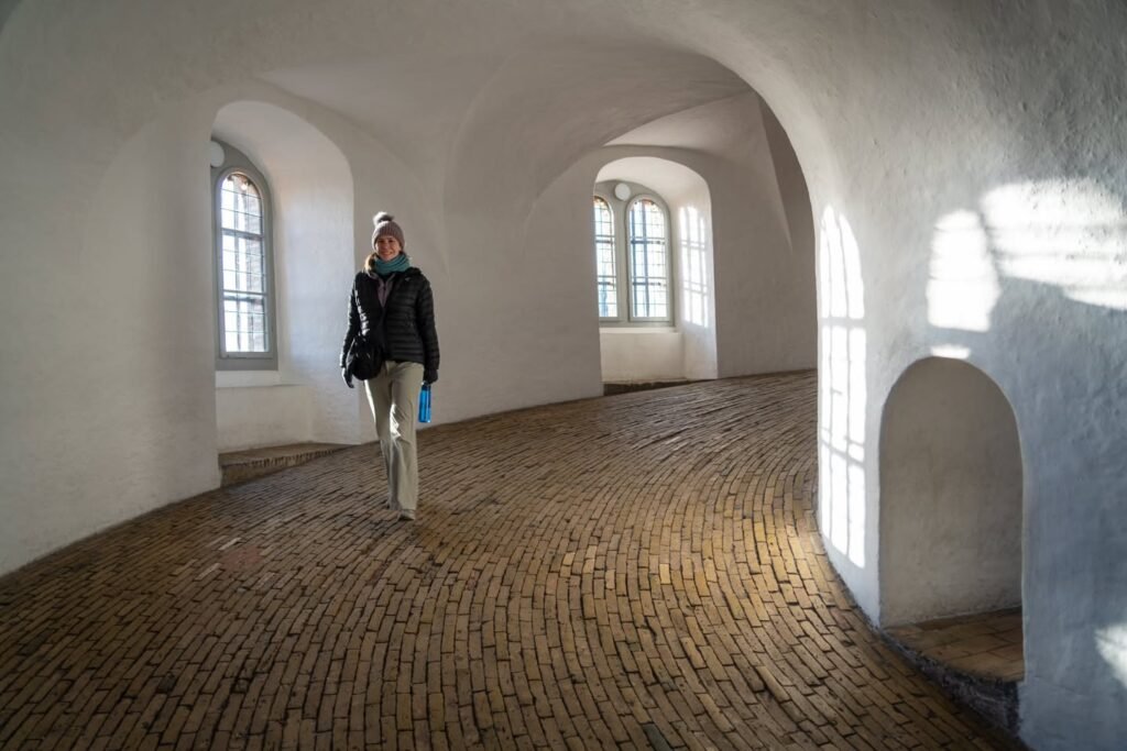 A person walks through a white arched room with a wooden floor. Large windows provide natural light in a historic space.