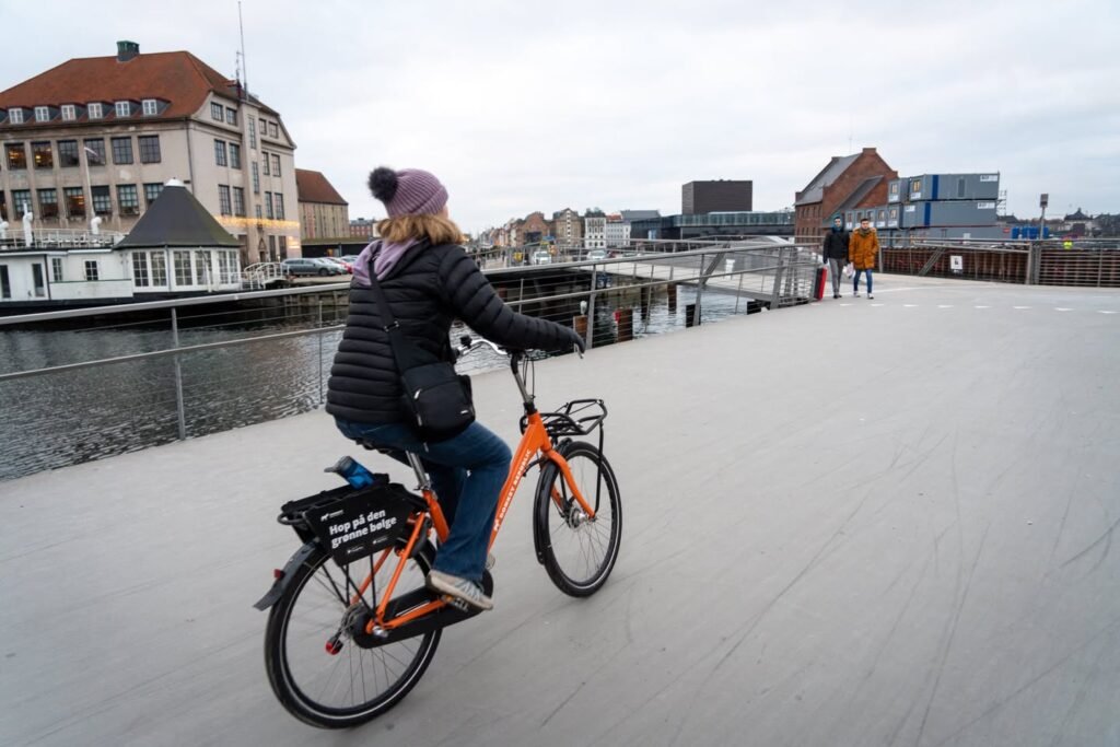 A person rides a bright orange bicycle along a waterfront promenade in Copenhagen. Historic European-style buildings line the harbor in the background under an overcast sky.