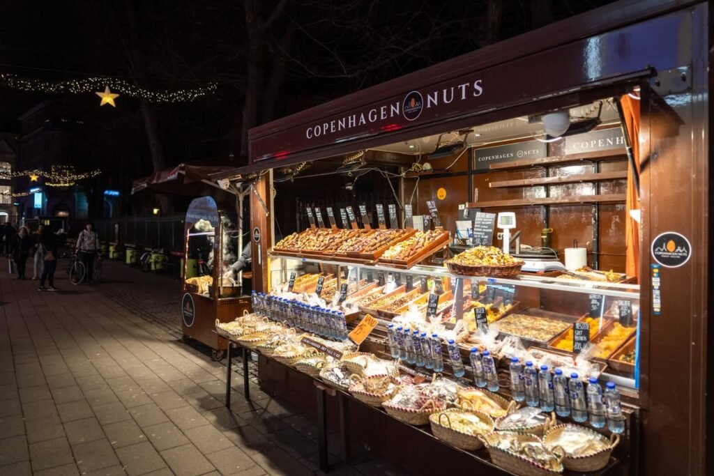 A brightly lit market stall in a European Christmas market displays an assortment of nuts and candies, topped by a sign reading 'Copenhagen Nuts.' Twinkling lights and a warm atmosphere add to the festive scene.