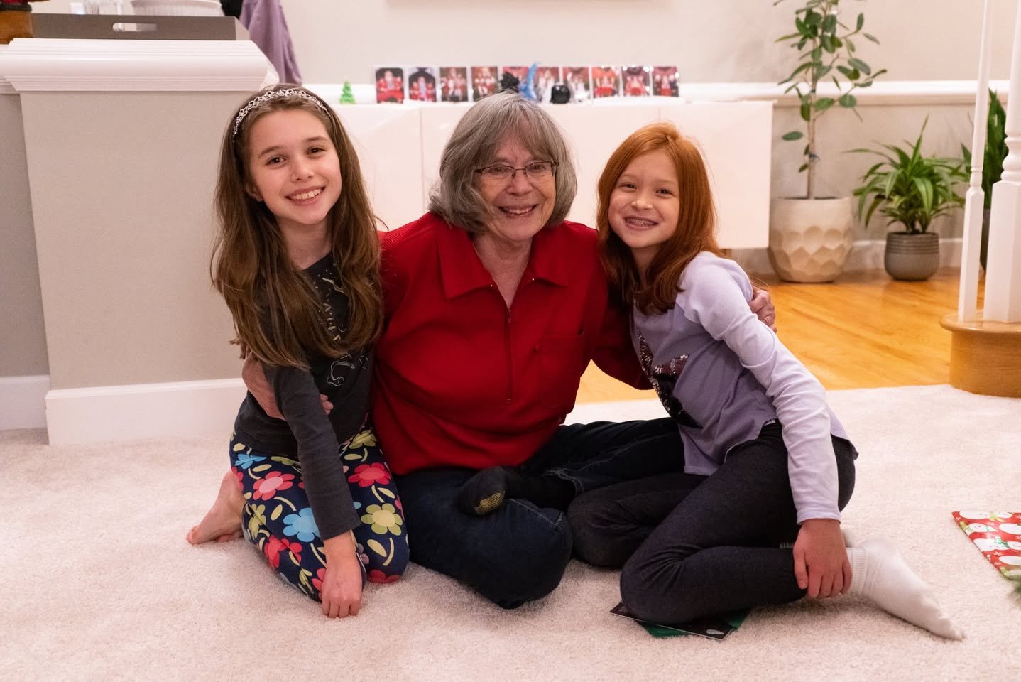 Close-up of a family of three sitting on a carpet, with the central figure wearing a vibrant red shirt. Background features household decor and plants.