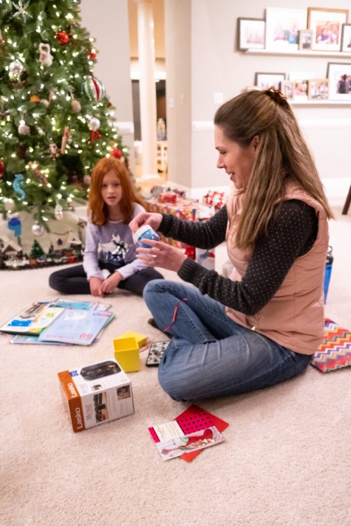 Two people sitting on the floor playing a board game in front of a decorated Christmas tree. The room has a warm, festive atmosphere with holiday ornaments.