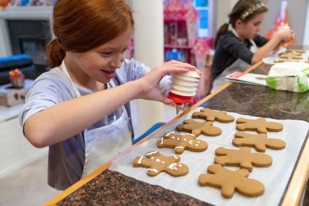 A girl carefully sprinkling white cookies on gingerbread figures with another child working in the background.