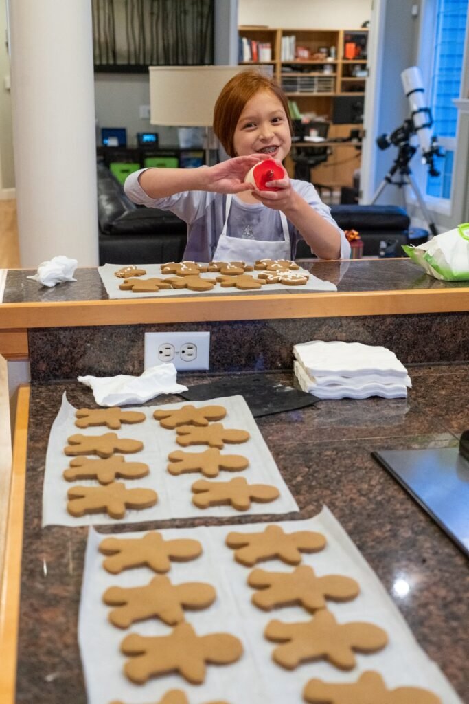 A child holds a red cookie cutter next to unfrosted gingerbread cookies on a kitchen counter.