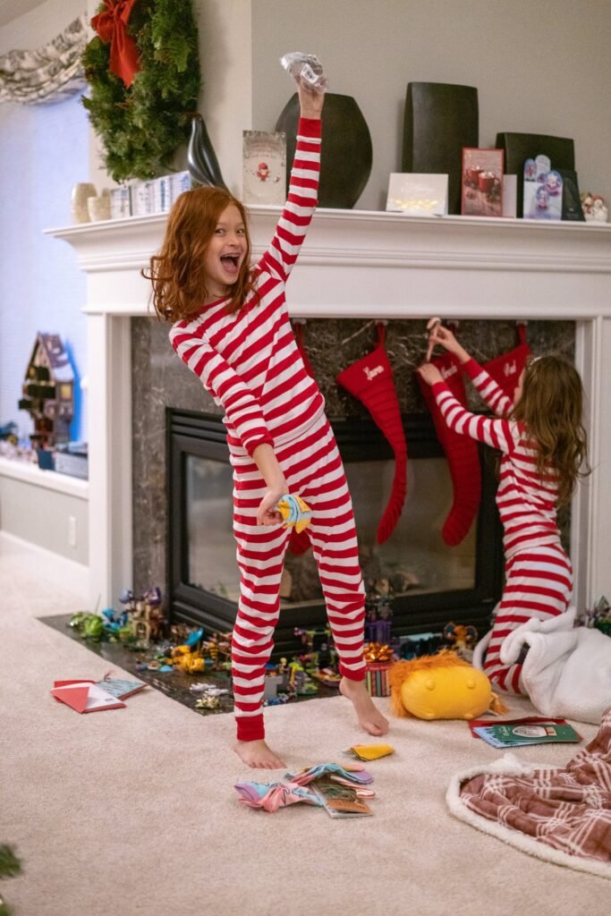 Children in matching red and white striped pajamas play near a fireplace. Red stockings hang from the mantel below a green wreath.