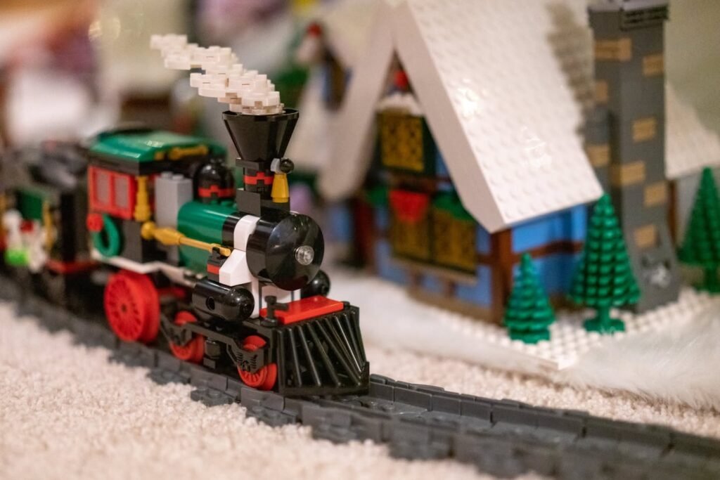 A bright green and red LEGO train travels on black tracks through a snowy village with small houses and trees. Four different close-up views showcase the festive scene.
