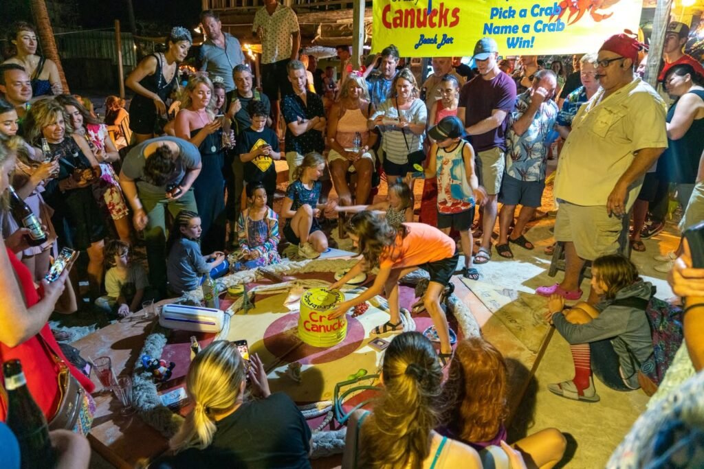 A large crowd of adults and children gathered around a sandy area for a crab-naming event at an outdoor party. A yellow banner in the background reads 'Canucks'.
