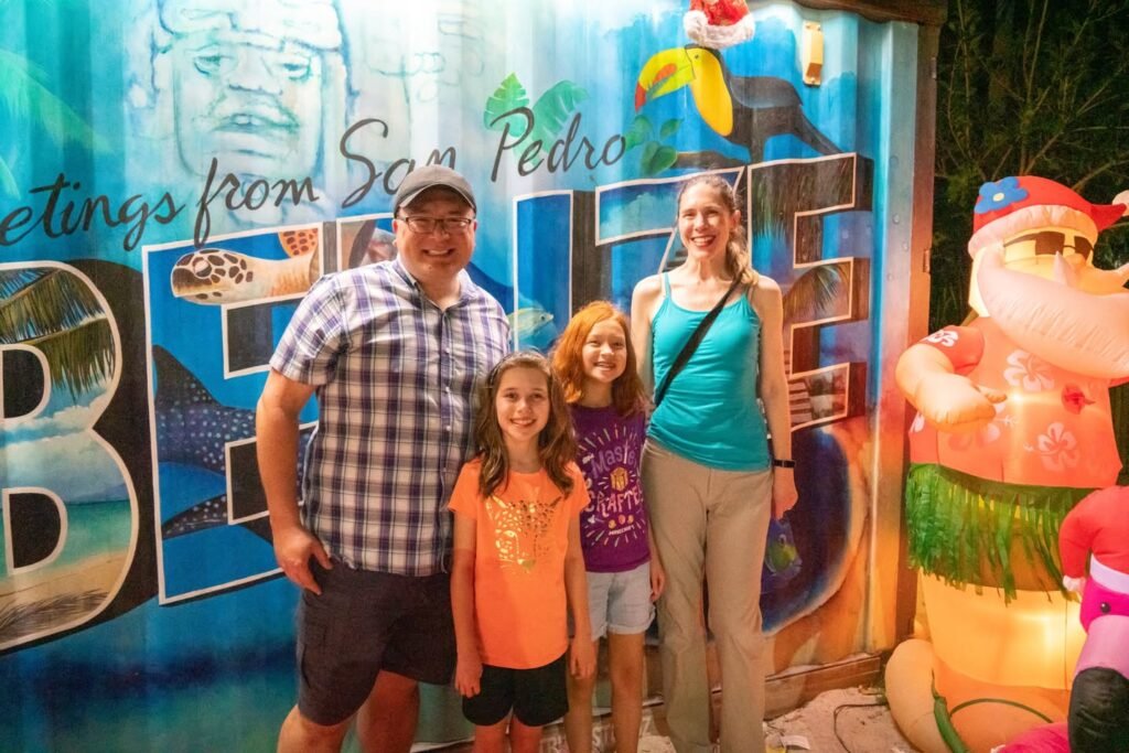 A family poses together in front of a colorful mural that reads 'Greetings from San Pedro.' The festive backdrop includes vibrant illustrations of a cartoon character and tropical elements.