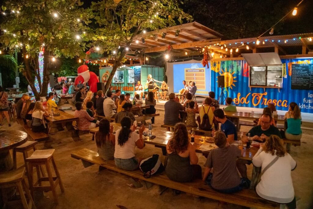 Patrons dine outdoors under string lights and trees at a lively evening event.