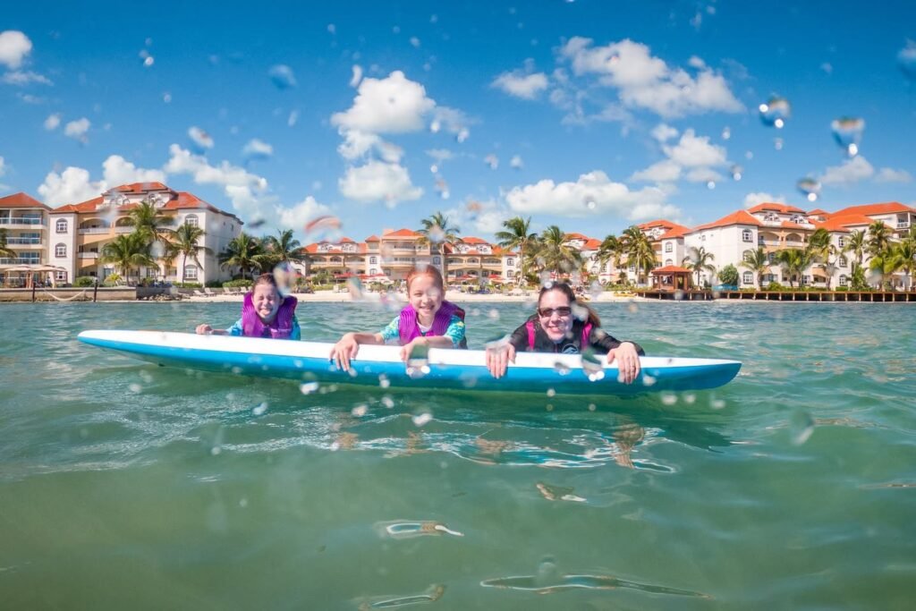 A group enjoys a wakeboard ride in clear waters. The backdrop features a scenic resort town with buildings and palm trees.