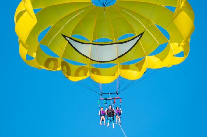 A yellow paraglider canopy with a painted smiley face design floats against a bright blue sky.