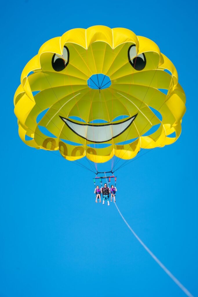 A yellow paraglider canopy with a painted smiley face design floats against a bright blue sky.
