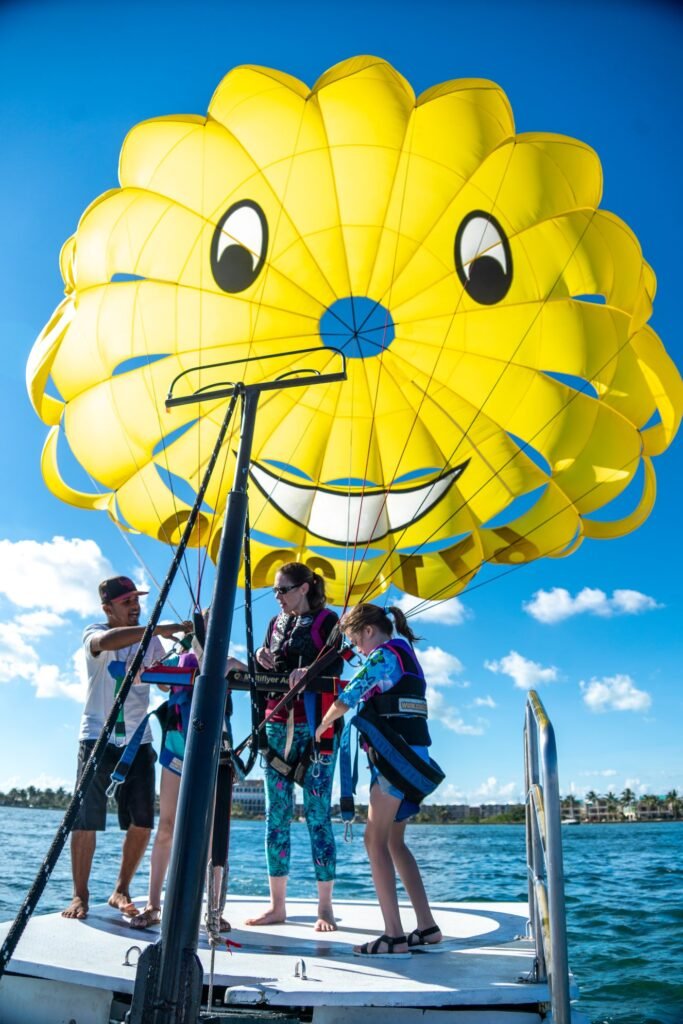 A large yellow parasail designed to look like a smiling face soars above two people seated on a boat in a tropical setting.