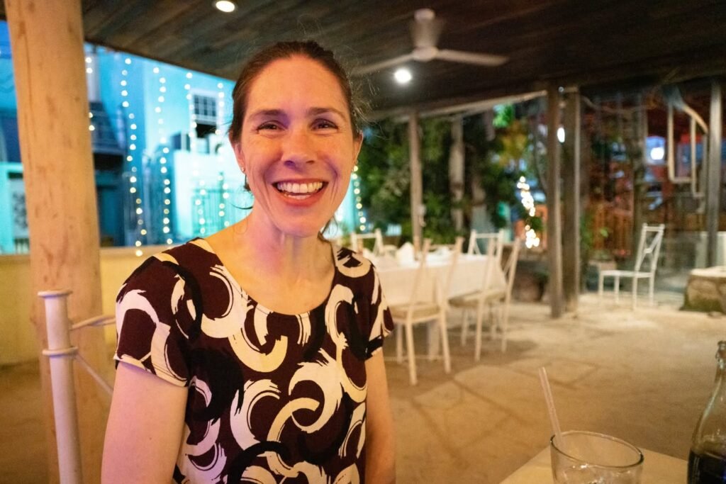 A person smiles warmly at an outdoor restaurant table with string lights in the background.