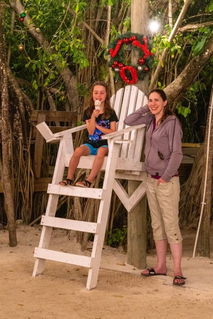 A person poses beside an oversized white lifeguard chair on a sandy beach under a tree decorated with a red Christmas wreath.