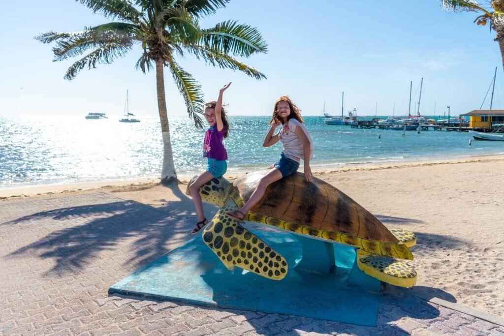 A child rides a large sea turtle statue on the beach near palm trees and the ocean. The sea turtle sculpture has a blue base and sits on the sand.