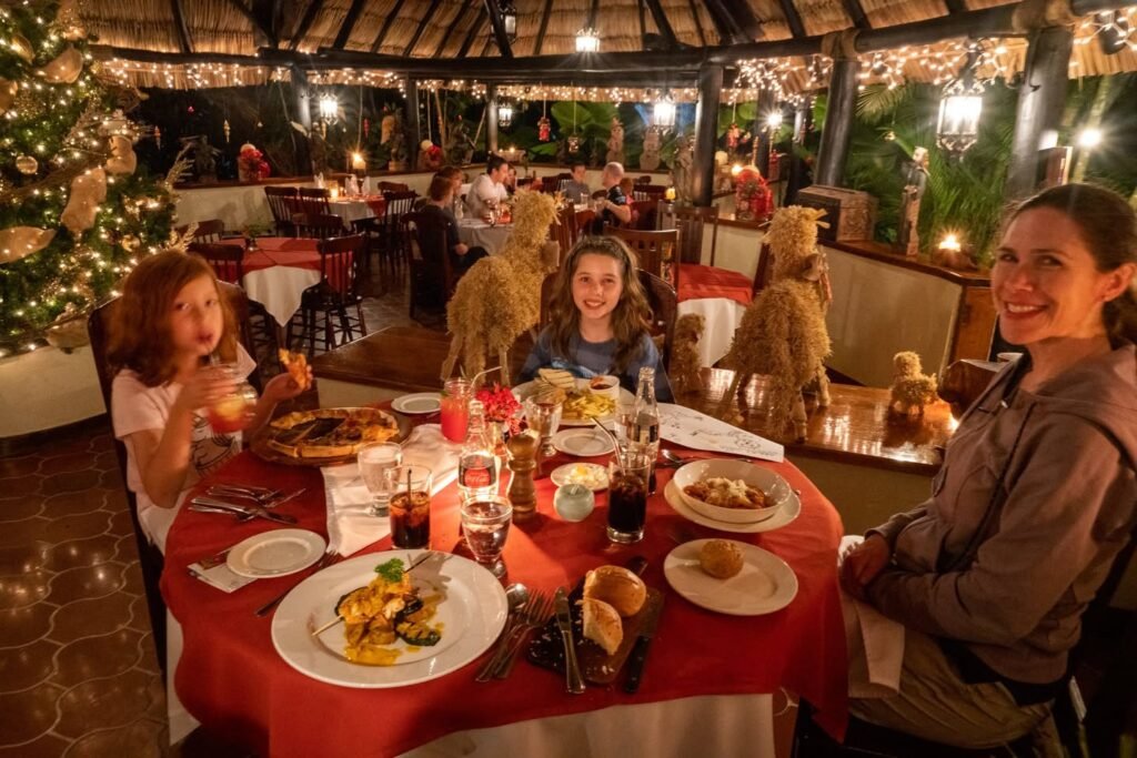 A family enjoys a festive meal at a table with a red tablecloth under warm holiday lighting.