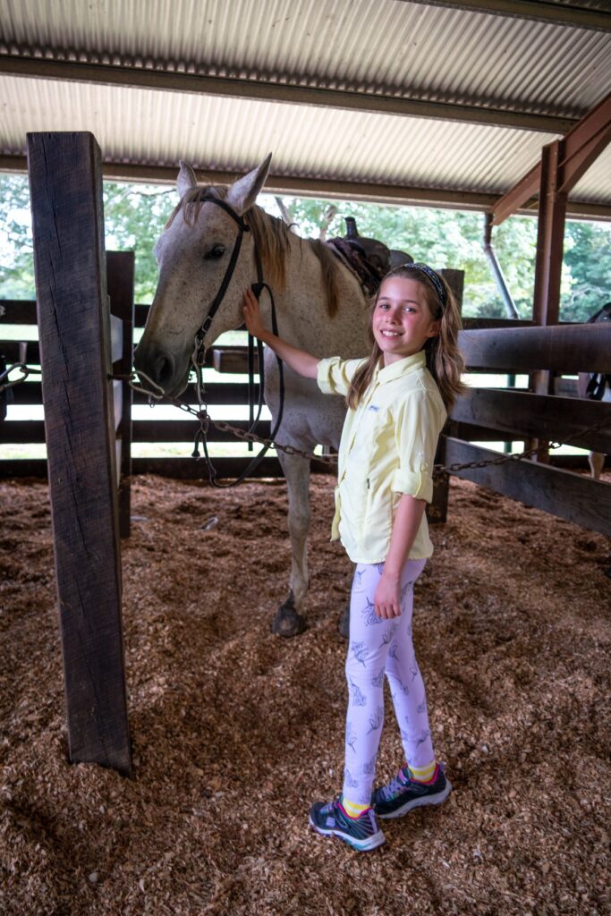 A child and a buckskin horse stand together in a rustic barn. The horse wears a bridle, and the ground is covered in wood chips.