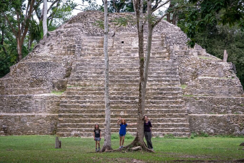 A family stands before the stepped stone pyramid at an ancient Mayan archaeological site. The dense green jungle surrounds the historic ruins.